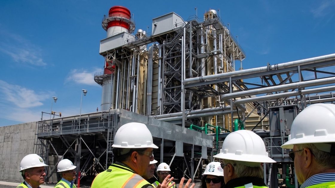 Equipe com capacetes diante de uma planta industrial com linhas de tubulação